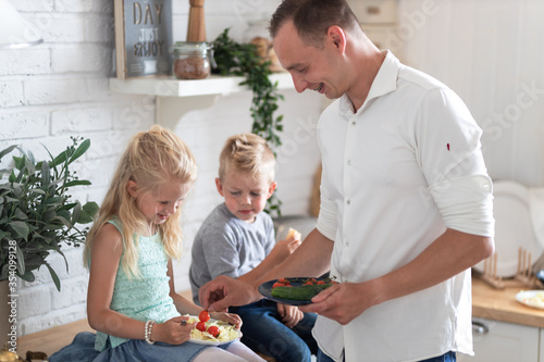 family father with three kids two sons and daughter eating healthy food in kitchen at home, dad puts green salad on plates to his kids.