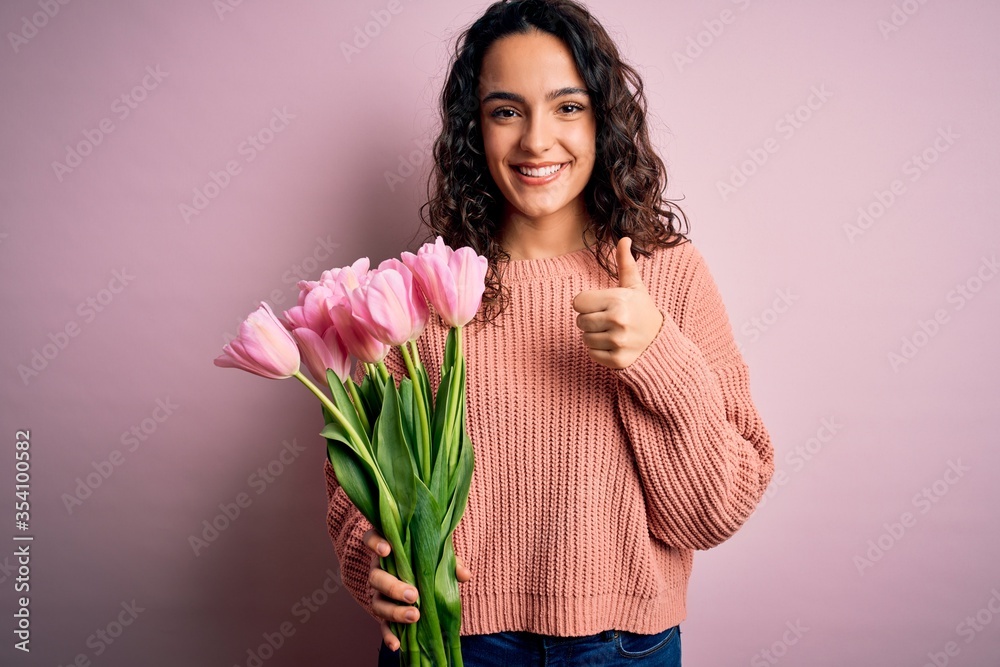 Young beautiful romantic woman with curly hair holding bouquet of pink tulips doing happy thumbs up gesture with hand. Approving expression looking at the camera showing success. Young beautiful romantic woman with curly hair holding bouquet of pink tulips doing happy thumbs up gesture with hand. Approving expression looking at the camera showing success.