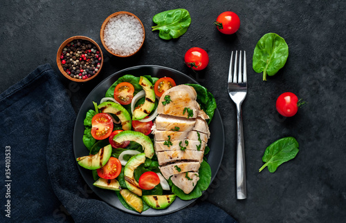  Grilled chicken breast and grilled avocado salad with cherry tomatoes, spinach, in a black plate on a stone background