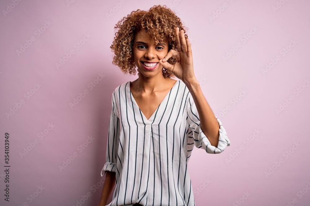 Beautiful african american woman with curly hair wearing striped t-shirt over pink background smiling positive doing ok sign with hand and fingers. Successful expression. Beautiful african american woman with curly hair wearing striped t-shirt over pink background smiling positive doing ok sign with hand and fingers. Successful expression.