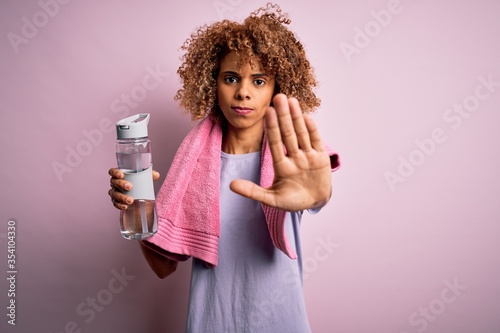 Young african american sportswoman doing sport wearing towel drinking bottle of water with open hand doing stop sign with serious and confident expression, defense gesture