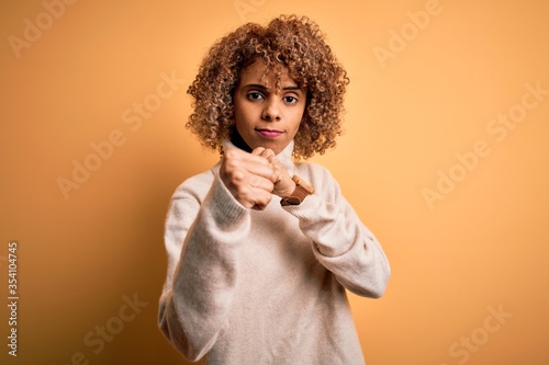 Young beautiful african american woman wearing turtleneck sweater over yellow background Ready to fight with fist defense gesture, angry and upset face, afraid of problem