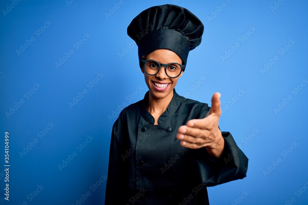 Young african american chef woman wearing cooker uniform and hat over blue background smiling friendly offering handshake as greeting and welcoming. Successful business. Young african american chef woman wearing cooker uniform and hat over blue background smiling friendly offering handshake as greeting and welcoming. Successful business.