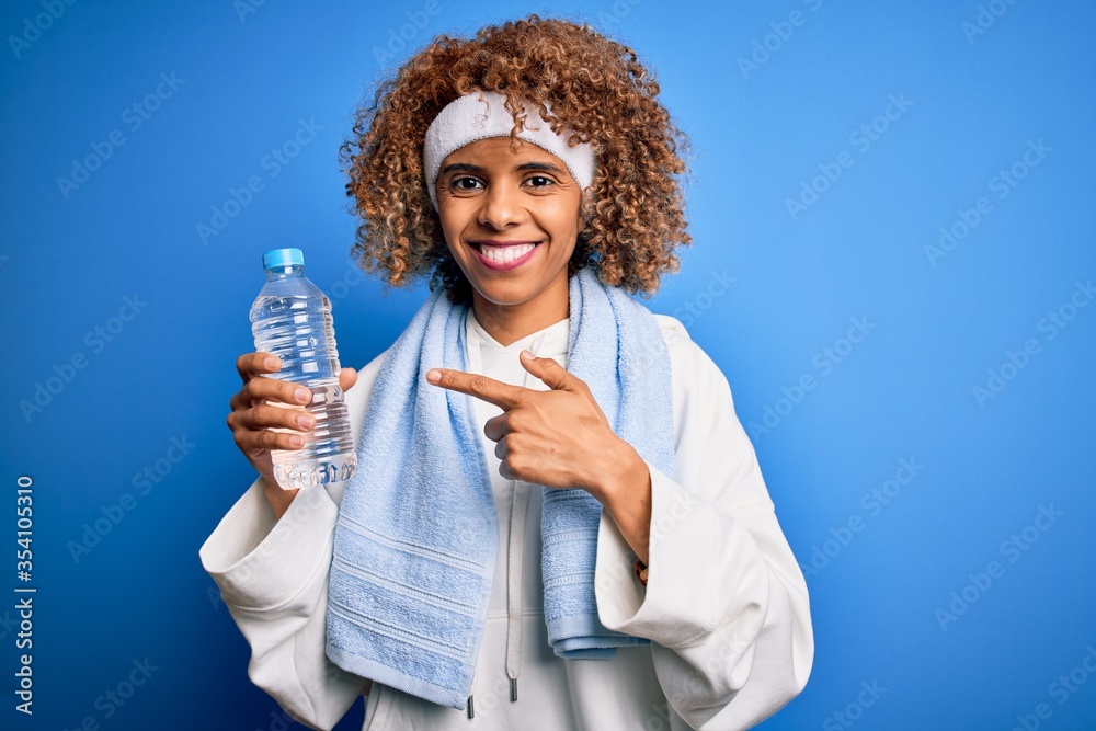 Young african american sportswoman doing sport wearing towel drinking botte of water very happy pointing with hand and finger Young african american sportswoman doing sport wearing towel drinking botte of water very happy pointing with hand and finger