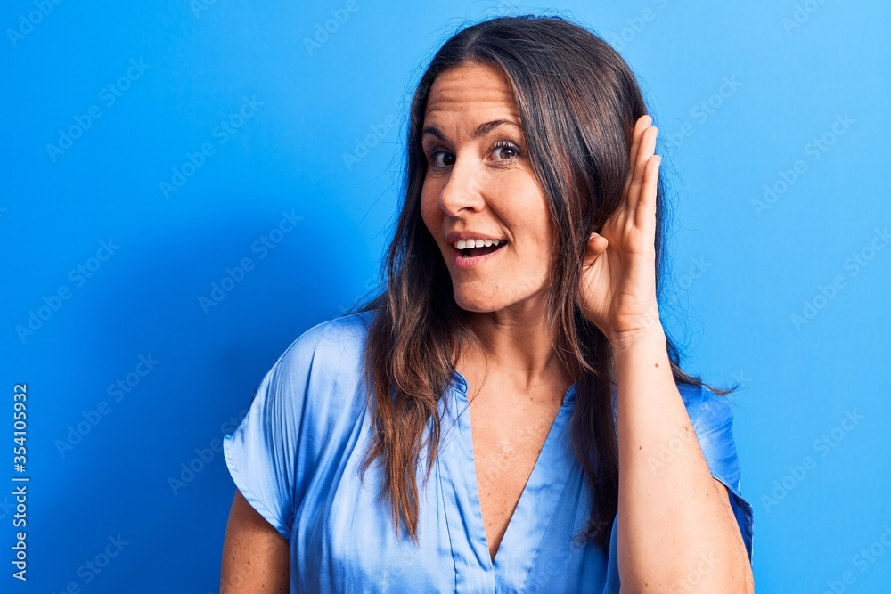 Young beautiful brunette woman wearing casual t-shirt standing over isolated blue background smiling with hand over ear listening and hearing to rumor or gossip. Deafness concept. Young beautiful brunette woman wearing casual t-shirt standing over isolated blue background smiling with hand over ear listening and hearing to rumor or gossip. Deafness concept.
