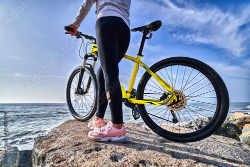 Fitness woman with bicycle stands on the stones on the seashore against the blue sky. Athletic healthy people with active and sporty lifestyle © Goffkein