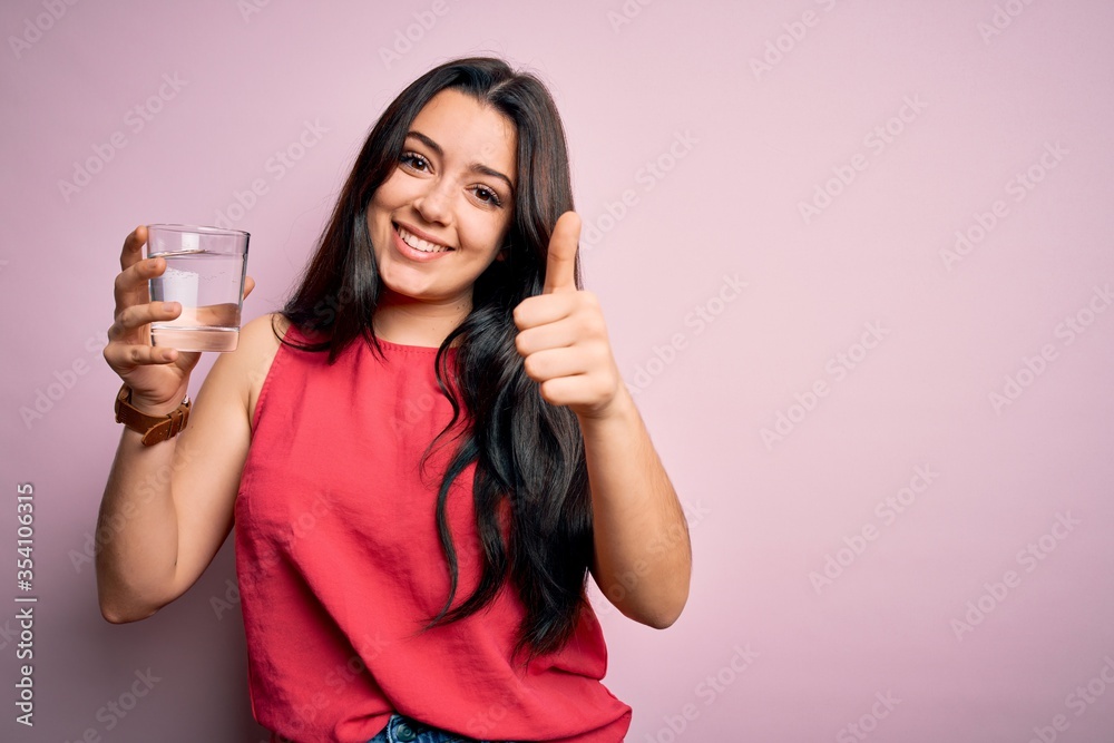 Young brunette woman drinking a glass of fresh water over pink isolated background happy with big smile doing ok sign, thumb up with fingers, excellent sign Young brunette woman drinking a glass of fresh water over pink isolated background happy with big smile doing ok sign, thumb up with fingers, excellent sign