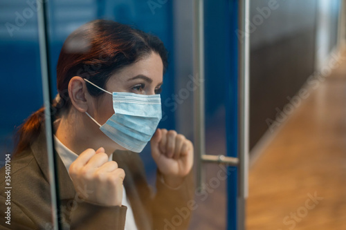 A girl in a medical mask is isolated due to biological hazard. The business woman in the suit is quarantined and leaned against the glass door with sadness. The epidemic of the virus.