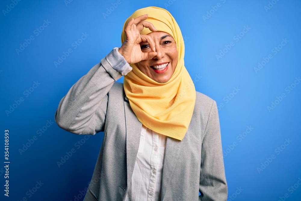 Middle age brunette business woman wearing muslim traditional hijab over blue background doing ok gesture with hand smiling, eye looking through fingers with happy face. Middle age brunette business woman wearing muslim traditional hijab over blue background doing ok gesture with hand smiling, eye looking through fingers with happy face.