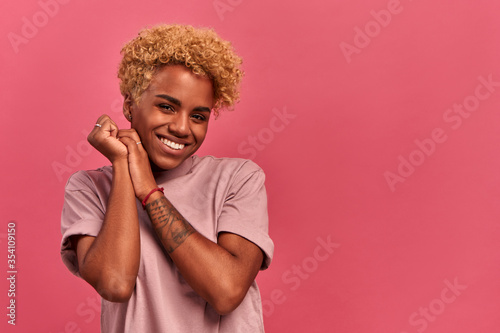 Happy cheerful african american woman rejoicing gift  keeps hands together near face  focused away  notices desirable things  laughs on a pink background. Personal offers and discounts in online store
