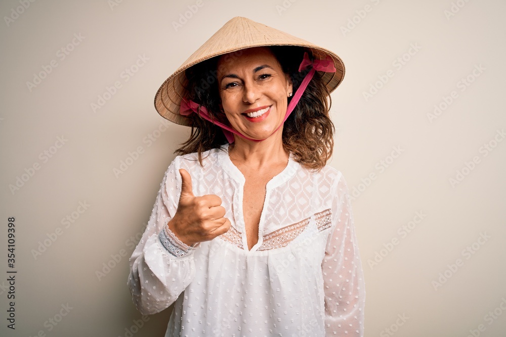Middle age brunette woman wearing asian traditional conical hat over white background doing happy thumbs up gesture with hand. Approving expression looking at the camera showing success. Middle age brunette woman wearing asian traditional conical hat over white background doing happy thumbs up gesture with hand. Approving expression looking at the camera showing success.