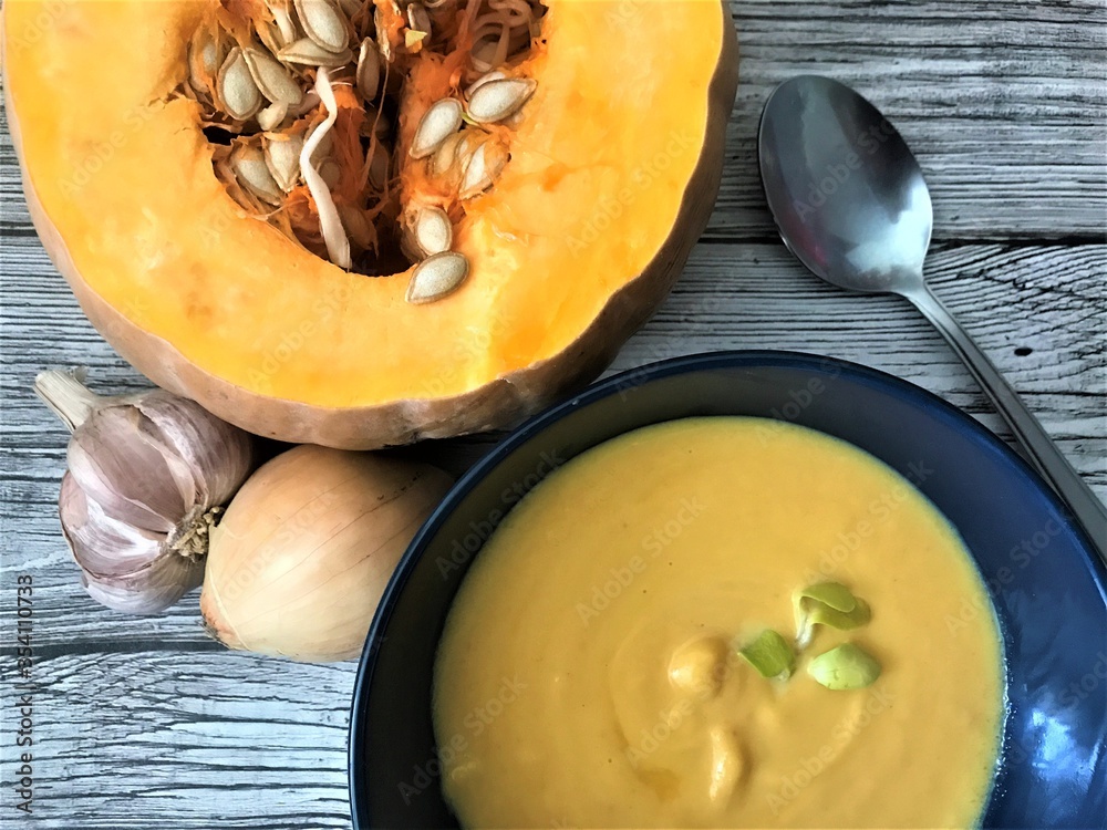 Dark blue bowl with pumpkin puree soup with a spoon on the right, untreated head of garlic and onion on the left and cut pumpkin with seeds on top against the background of old gray wood
Dark blue bowl with pumpkin puree soup with a spoon on the right, untreated head of garlic and onion on the left and cut pumpkin with seeds on top against the background of old gray wood