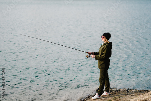 Fisherman with rod, spinning reel on river bank. Sunset. Fishing for pike, perch, carp. Woman catching fish, pulling rod while fishing at weekend. Girl fishing from beach lake or pond with text space.