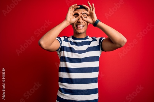 Handsome african american man wearing casual striped t-shirt standing over red background doing ok gesture like binoculars sticking tongue out, eyes looking through fingers. Crazy expression.