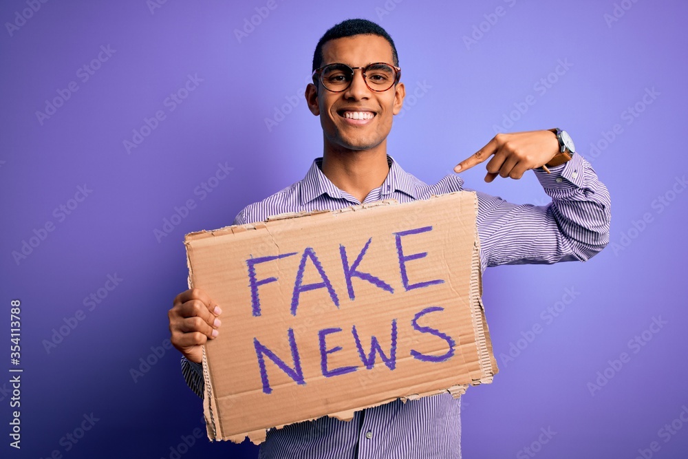 Young handsome african american man protesting holding banner with fake news message with surprise face pointing finger to himself Young handsome african american man protesting holding banner with fake news message with surprise face pointing finger to himself