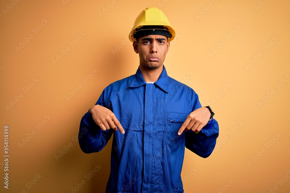 Young handsome african american worker man wearing blue uniform and security helmet Pointing down looking sad and upset, indicating direction with fingers, unhappy and depressed.
