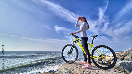 Fitness woman with a bicycle stands on the stones on the seashore and looks into the distance on a clear sunny day. Athletic healthy people with sporty and active lifestyle photo