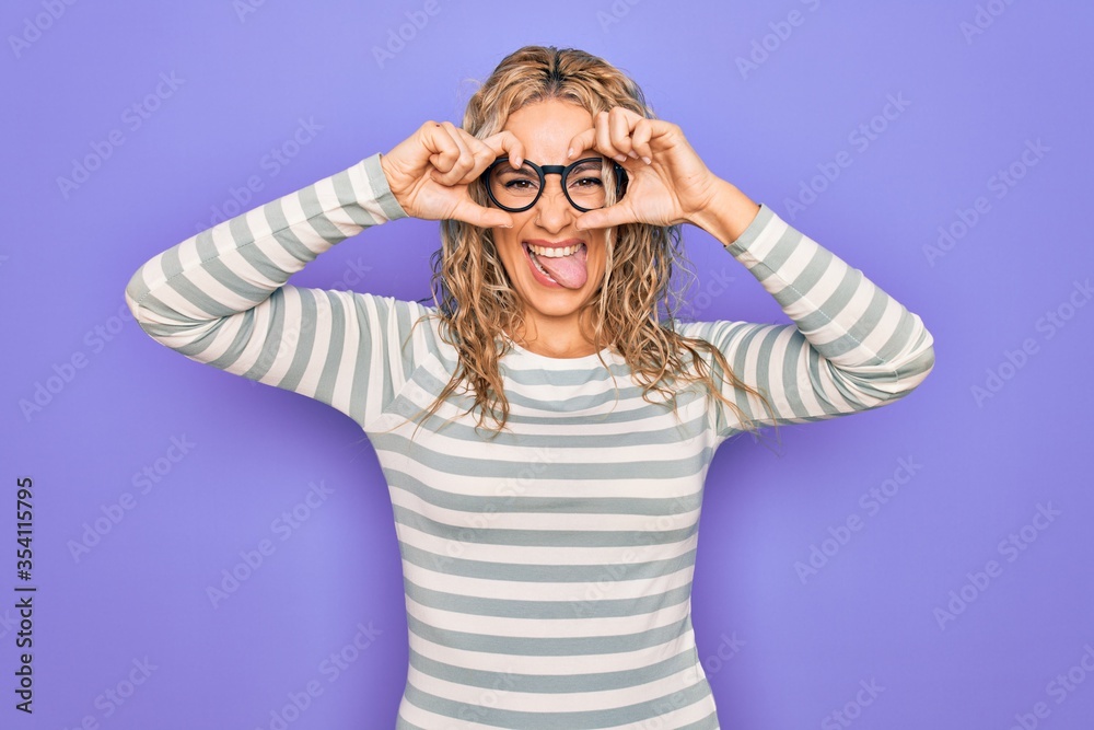 Beautiful blonde woman wearing casual striped t-shirt and glasses over purple background doing ok gesture like binoculars sticking tongue out, eyes looking through fingers. Crazy expression. Beautiful blonde woman wearing casual striped t-shirt and glasses over purple background doing ok gesture like binoculars sticking tongue out, eyes looking through fingers. Crazy expression.