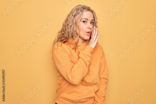 Young beautiful blonde sporty woman wearing casual sweatshirt over yellow background hand on mouth telling secret rumor, whispering malicious talk conversation