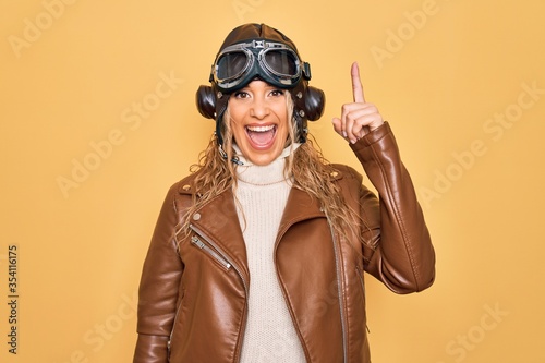 Obraz na płótnie Young beautiful blonde aviator woman wearing vintage pilot helmet whit glasses and jacket pointing finger up with successful idea