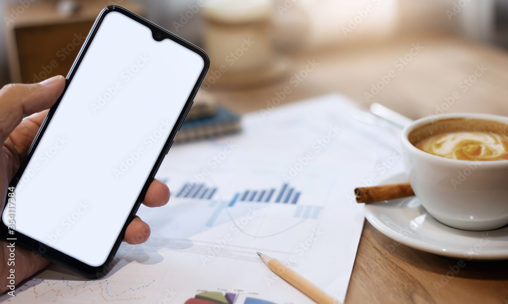Close up of hand holding mobile phone with paperwork of investment and coffee drink on wooden table Close up of hand holding mobile phone with paperwork of investment and coffee drink on wooden table