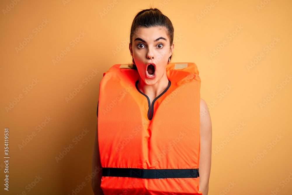 Young beautiful brunette woman wearing orange safe lifejacket over yellow background afraid and shocked with surprise expression, fear and excited face. Young beautiful brunette woman wearing orange safe lifejacket over yellow background afraid and shocked with surprise expression, fear and excited face.