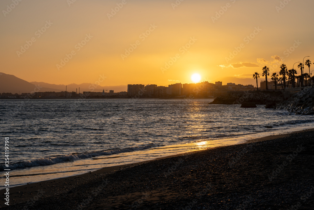Summer sunset. Orange sky over the sea, beach with palm trees, city on the horizon.