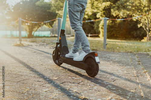 Person fährt entlang auf der Straße im Park einen E-Scooter bei Sonnenuntergang. Junger Mensch fährt einen elektrischen Cityroller im Sommer mit Hilfe neuster Technologie.