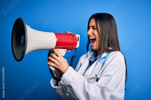 Hispanic doctor woman wearing medical white coat shouting angry on protest through megaphone. Yelling excited on ludspeaker talking and screaming news