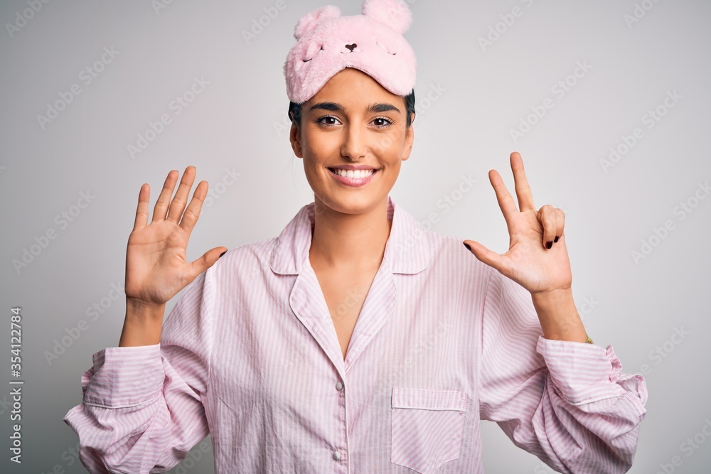 Young beautiful brunette woman wearing pajama and sleep mask over white background showing and pointing up with fingers number eight while smiling confident and happy. Young beautiful brunette woman wearing pajama and sleep mask over white background showing and pointing up with fingers number eight while smiling confident and happy.
