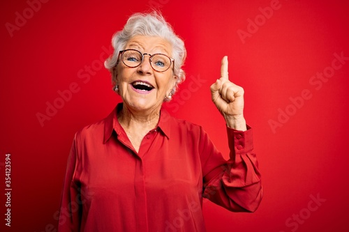 Senior beautiful grey-haired woman wearing casual shirt and glasses over red background pointing finger up with successful idea. Exited and happy. Number one.