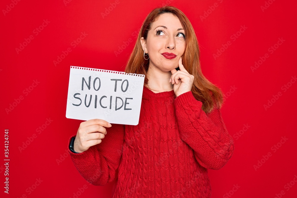 Beautiful redhead woman holding banner showing no to suicide message over red background serious face thinking about question, very confused idea Beautiful redhead woman holding banner showing no to suicide message over red background serious face thinking about question, very confused idea