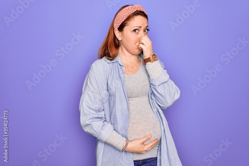 Young beautiful redhead pregnant woman expecting baby over isolated purple background smelling something stinky and disgusting, intolerable smell, holding breath with fingers on nose. Bad smell