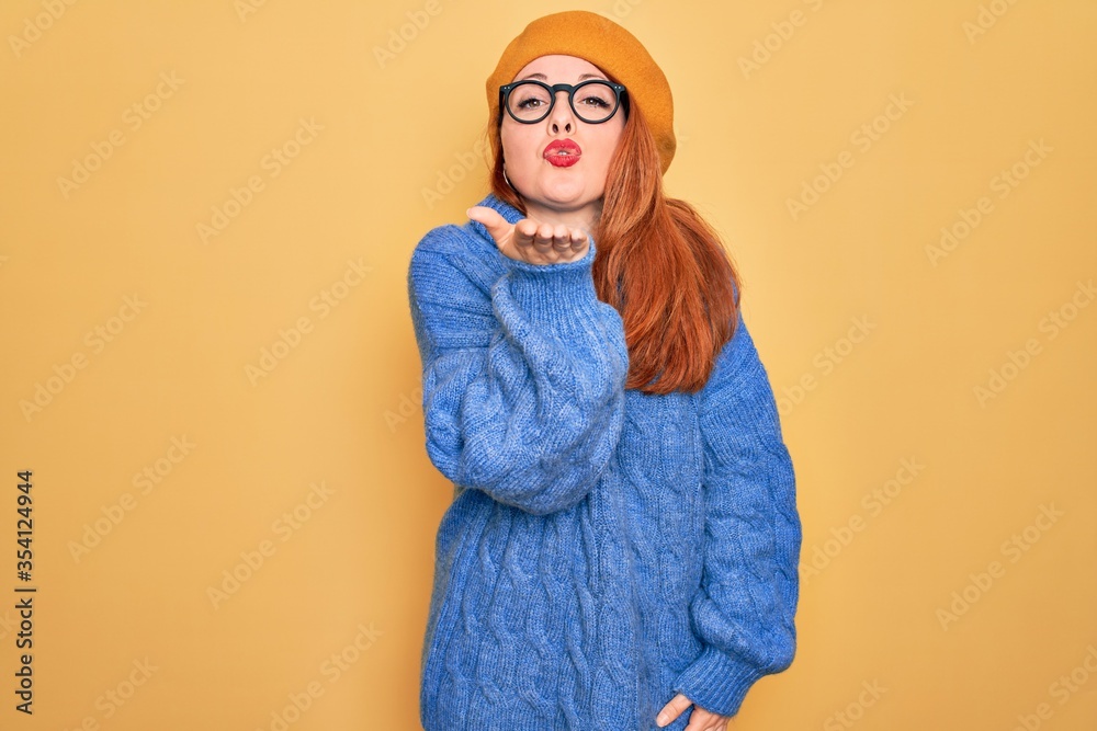 Young beautiful redhead woman wearing french beret and glasses over yellow background looking at the camera blowing a kiss with hand on air being lovely and sexy. Love expression.