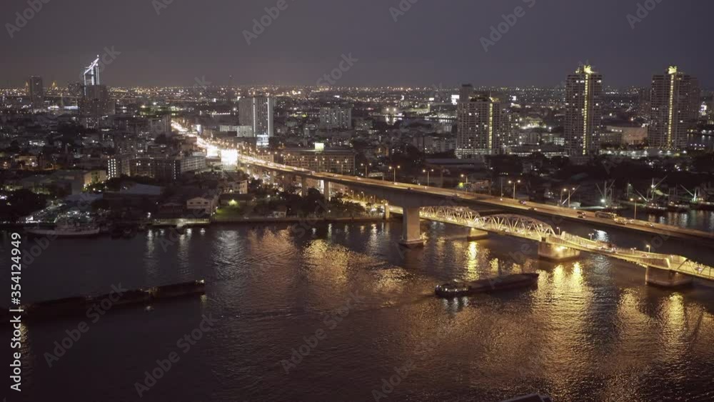 Lockdown shot of vehicles on bridge over river by illuminated buildings in modern city against sky at night - Bangkok, Thailand Lockdown shot of vehicles on bridge over river by illuminated buildings in modern city against sky at night - Bangkok, Thailand