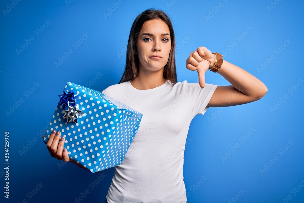 Young beautiful brunette woman holding birthday gift over isolated blue background with angry face, negative sign showing dislike with thumbs down, rejection concept Young beautiful brunette woman holding birthday gift over isolated blue background with angry face, negative sign showing dislike with thumbs down, rejection concept