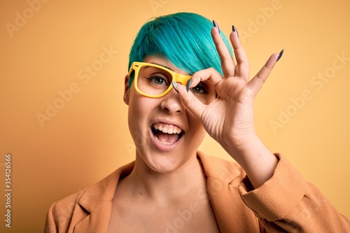 Young beautiful woman with blue fashion hair wearing casual glasses over yellow background with happy face smiling doing ok sign with hand on eye looking through fingers
