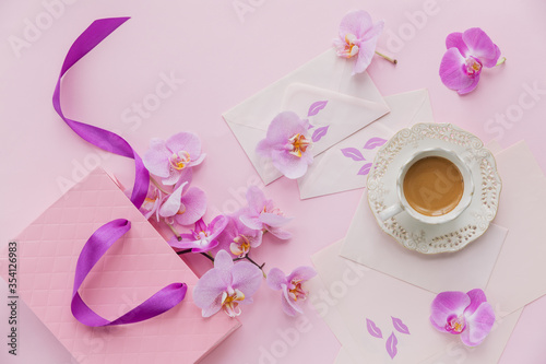 Delicate flatlay composition with morning cup of coffee with milk or cappuccino, letters, pink gift bag and orchid flowers on light pink background.