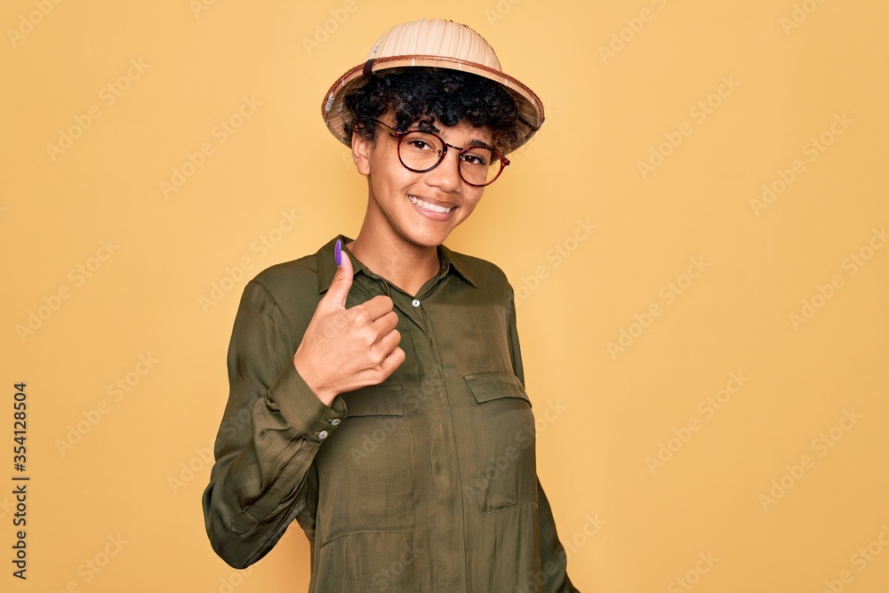 Young beautiful african american tourist woman wearing glasses and explorer hat doing happy thumbs up gesture with hand. Approving expression looking at the camera showing success. Young beautiful african american tourist woman wearing glasses and explorer hat doing happy thumbs up gesture with hand. Approving expression looking at the camera showing success.