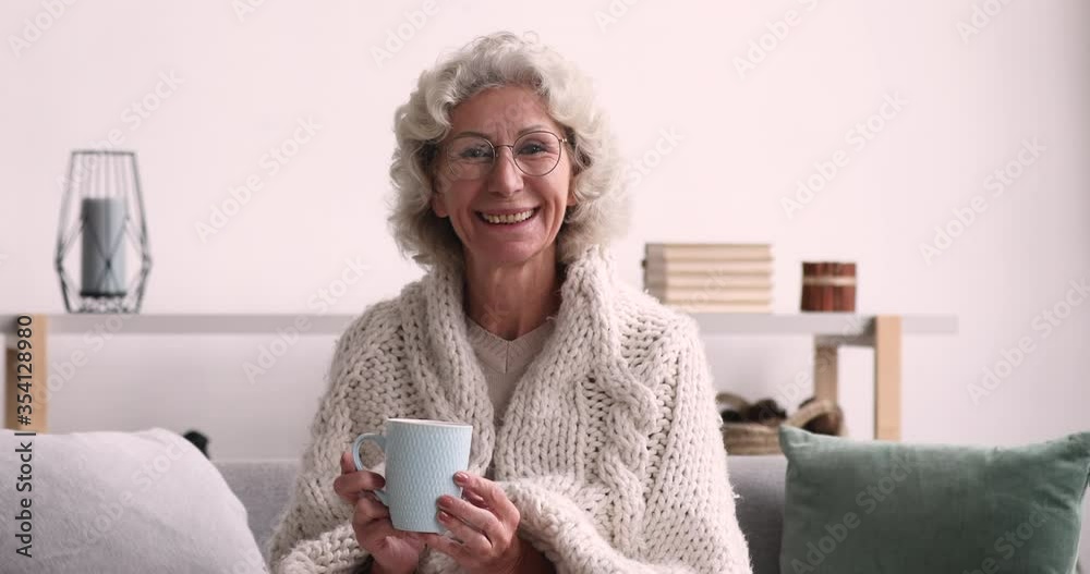 Peaceful happy middle aged mature woman in glasses looking at camera, sitting on comfortable couch covered in plaid with cup of hot tea or coffee in hands, enjoying cold winter season alone indoors. Peaceful happy middle aged mature woman in glasses looking at camera, sitting on comfortable couch covered in plaid with cup of hot tea or coffee in hands, enjoying cold winter season alone indoors.
