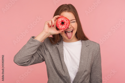 Health care and nutrition. Portrait of cheerful businesswoman in suit jacket looking through sweet doughnut, having fun with pastry, peeking into donut and smiling. indoor studio shot, pink background