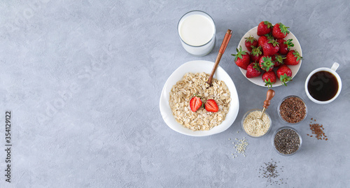 Ingredients for healthy breakfast concept banner. Bowl of oatmeal porridge, strawberries, milk and seeds on stone background with copy space