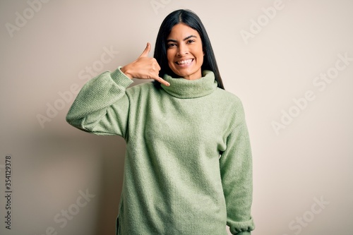 Young beautiful hispanic woman wearing green winter sweater over isolated background smiling doing phone gesture with hand and fingers like talking on the telephone. Communicating concepts. © Krakenimages.com