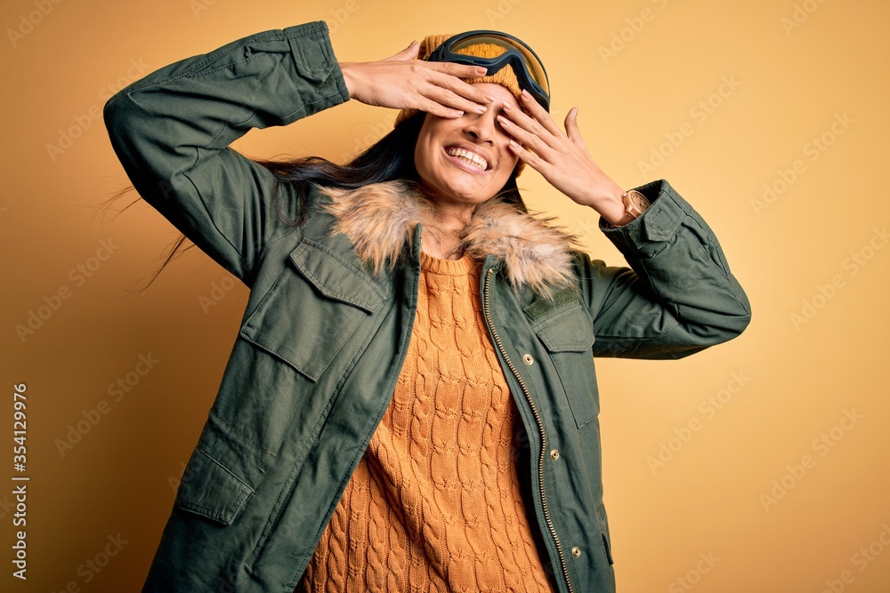 Young beautiful hispanic woman wearing ski glasses and coat for winter weather covering eyes with hands smiling cheerful and funny. Blind concept.
