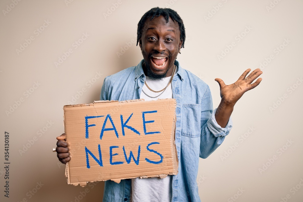 Young african american man holding banner with fake news message over white background very happy and excited, winner expression celebrating victory screaming with big smile and raised hands Young african american man holding banner with fake news message over white background very happy and excited, winner expression celebrating victory screaming with big smile and raised hands