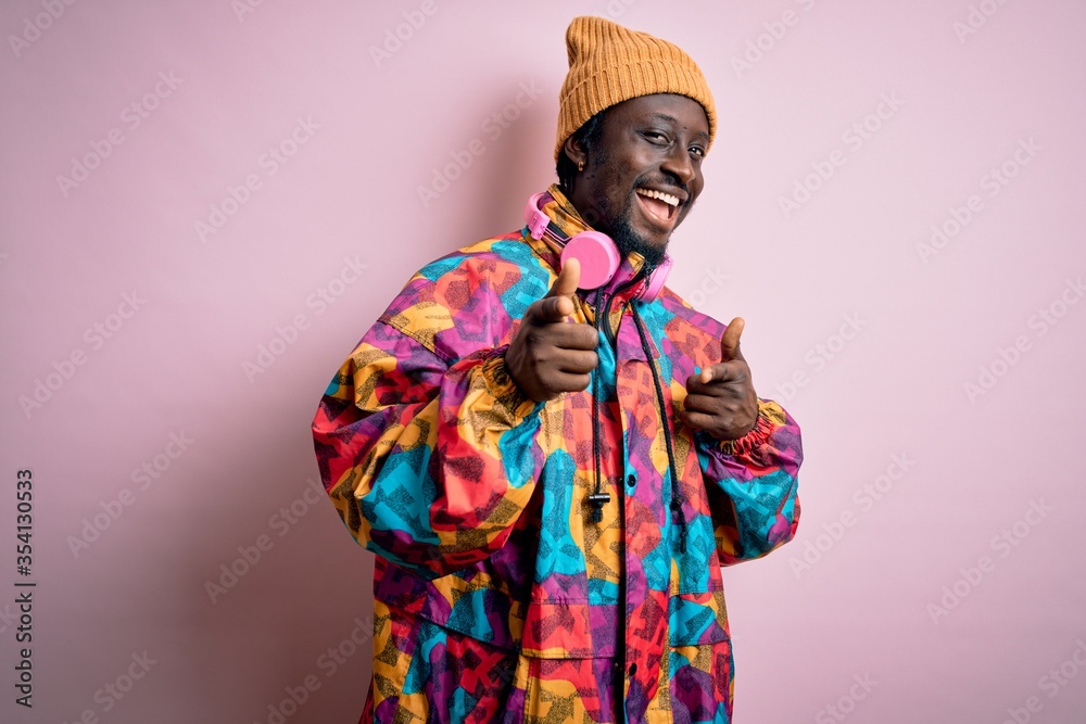 Young handsome african american man wearing colorful coat and cap over pink background pointing fingers to camera with happy and funny face. Good energy and vibes.
