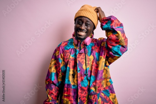 Young handsome african american man wearing colorful coat and cap over pink background confuse and wonder about question. Uncertain with doubt, thinking with hand on head. Pensive concept.