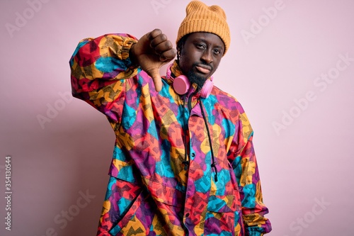 Young handsome african american man wearing colorful coat and cap over pink background looking unhappy and angry showing rejection and negative with thumbs down gesture. Bad expression.