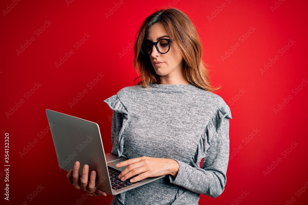 Naklejka premium Young beautiful brunette businesswoman wearing glasses using laptop over red background with a confident expression on smart face thinking serious