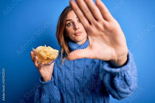 Young beautiful brunette woman holding bowl with potato chips over blue background with open hand doing stop sign with serious and confident expression  defense gesture
